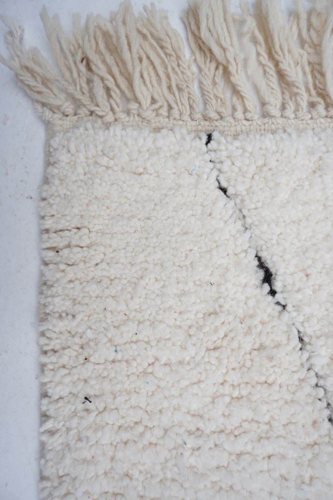 Tapis berbère doux en laine, couleur crème avec des motifs noirs géométriques et des franges délicates.