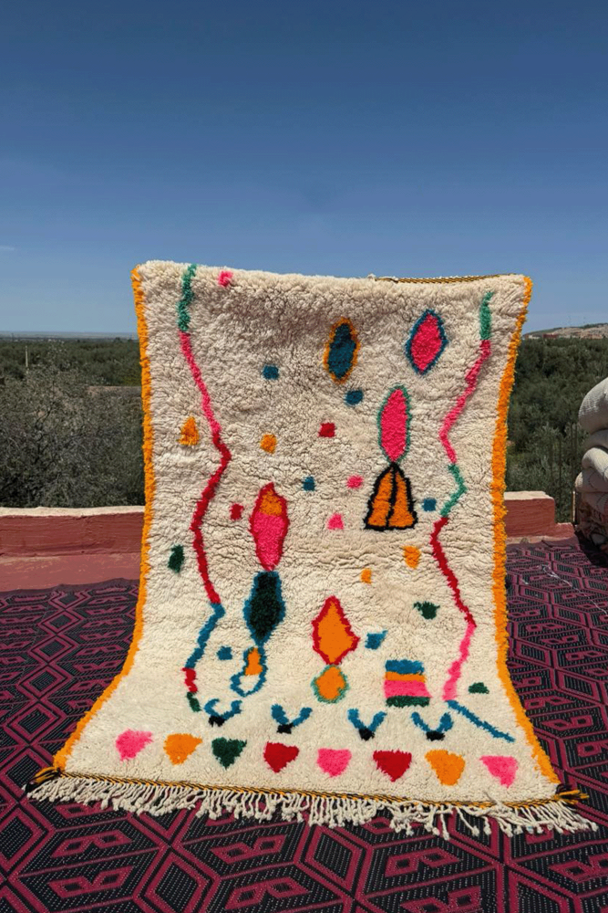 Tapiz bereber Azilal blanco con rombos y zigzags en rosa, verde y azul, con borde amarillo.