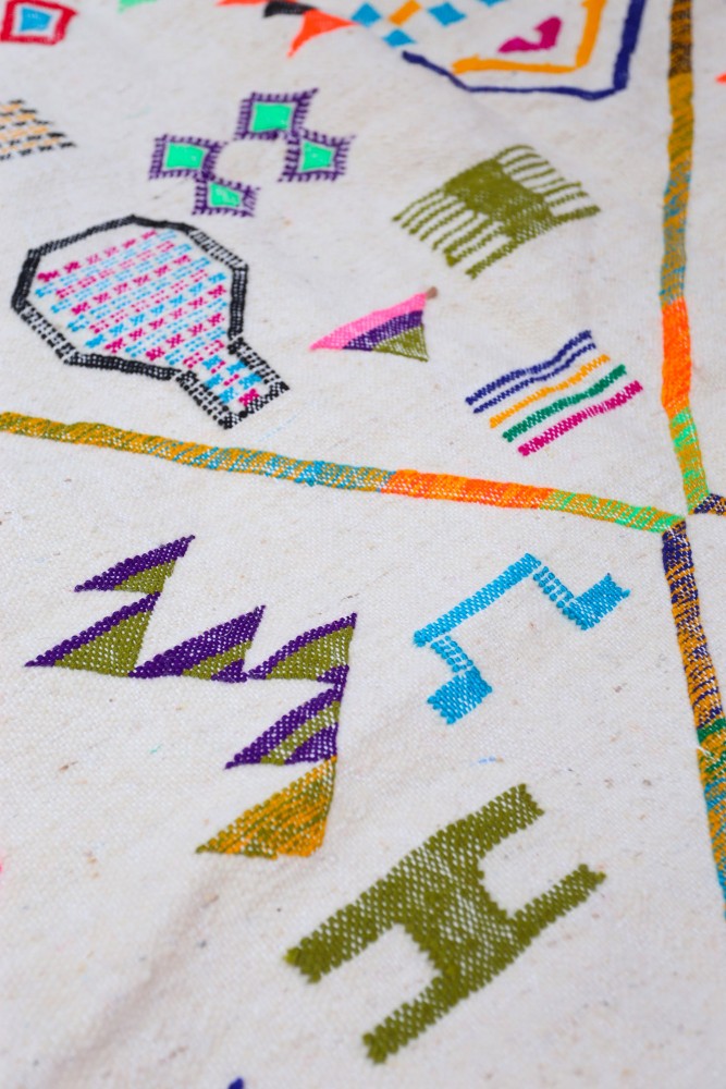 Tapis berbère coloré avec des motifs géométriques vifs et des teintes dominantes de beige, vert et violet.