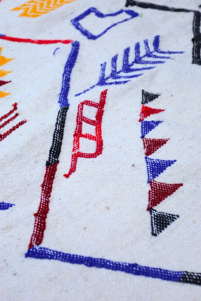 Tapis berbère coloré aux motifs géométriques en rouge, bleu, jaune et noir, sur fond blanc.