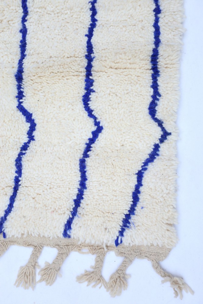 Tapis berbère en laine beige avec des motifs ondulés bleus et des franges, offrant une touche artisanale et chaleureuse.