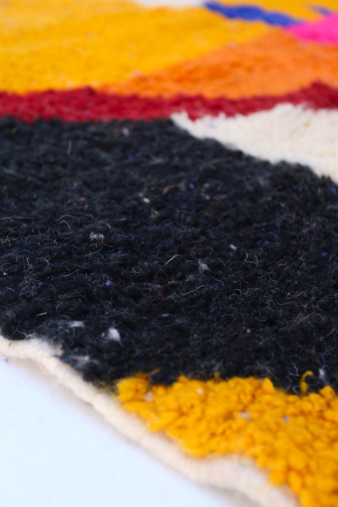 Tapis berbère coloré avec des motifs géométriques en noir, jaune, rouge et rose sur fond beige.