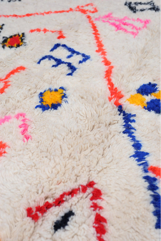 Tapis berbère à fond crème avec motifs colorés en bleu, rose et jaune.