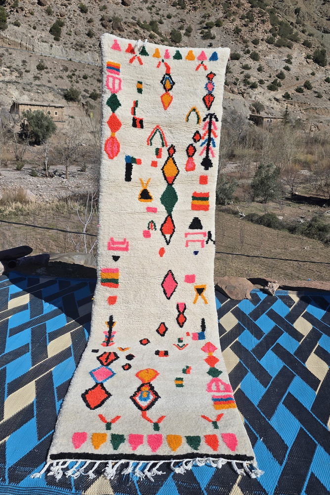 Tapis berbère aux motifs géométriques colorés, avec des teintes vives de rose, orange, noir et vert sur un fond crème.