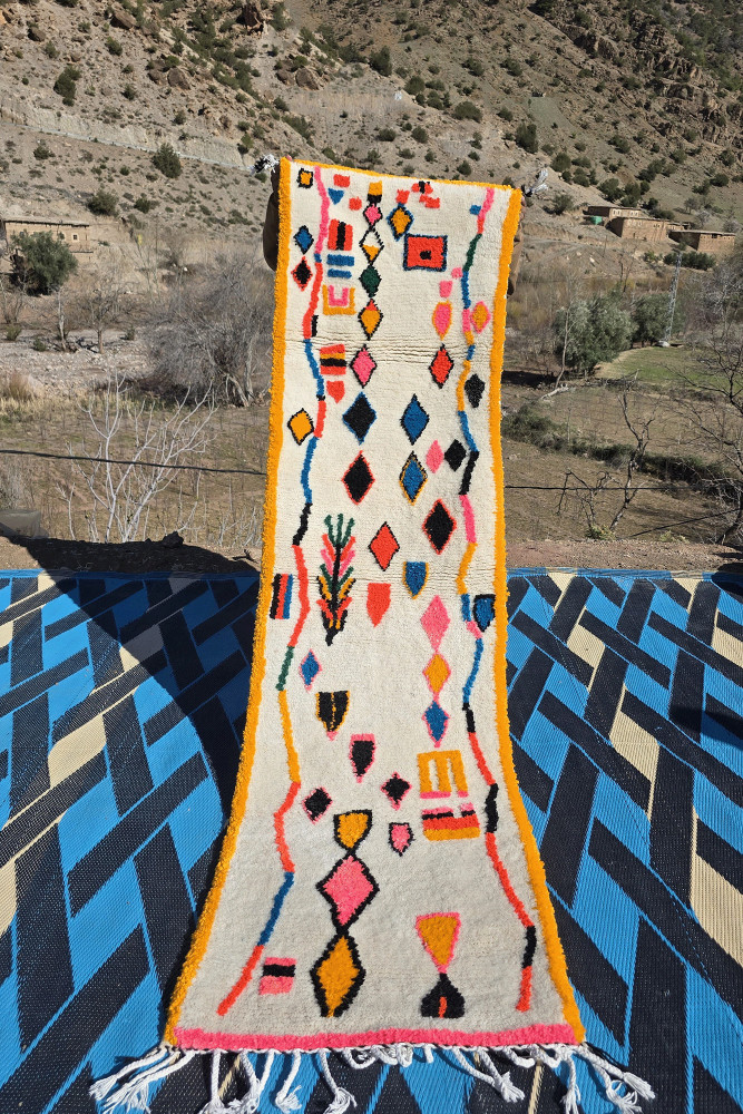 Tapis berbère aux motifs géométriques colorés, dominé par le blanc, le noir, le rose et l\'orange.