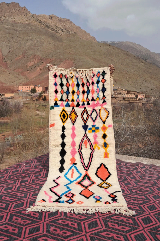 Tapis berbère coloré avec des motifs géométriques en rose, noir, orange et bleu sur fond crémeux.