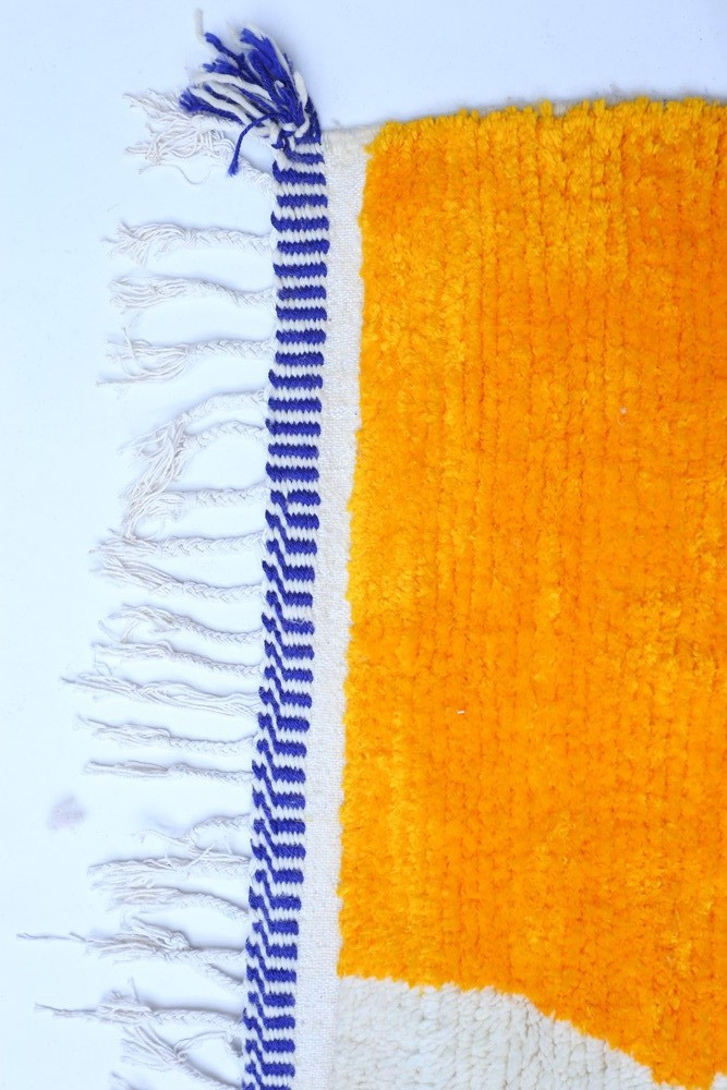 Tapis berbère aux motifs géométriques, couleurs jaune vif et blanc avec des franges bleues et blanches.