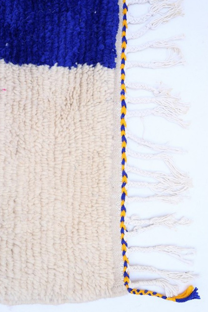 Tapis berbère en laine, avec des motifs géométriques, dominé par du bleu vif et du beige, orné de franges tressées jaunes.