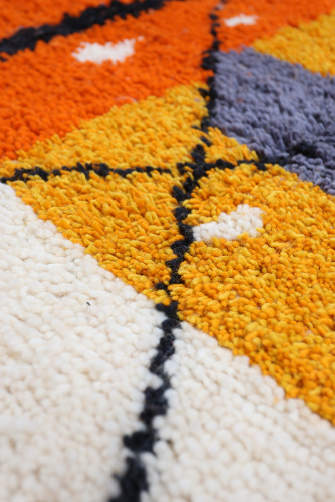 Tapis berbère aux motifs géométriques, dominant des teintes orange, jaune et gris avec des accents noirs et blancs.