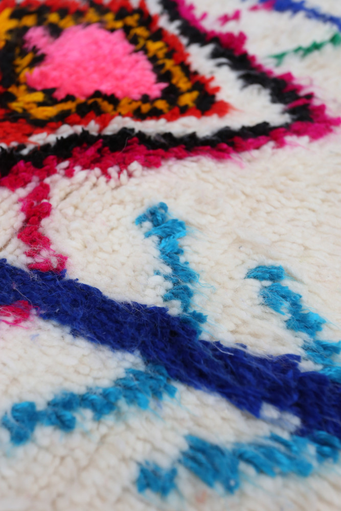 Tapis berbère aux motifs géométriques colorés, dominé par des teintes de rose, bleu et noir sur fond crème.