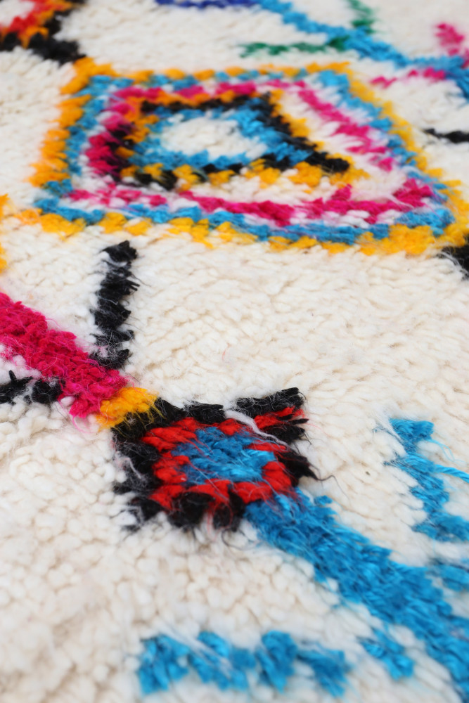 Tapis berbère aux motifs géométriques colorés, mêlant le blanc, le noir, le bleu, le rose et le jaune.
