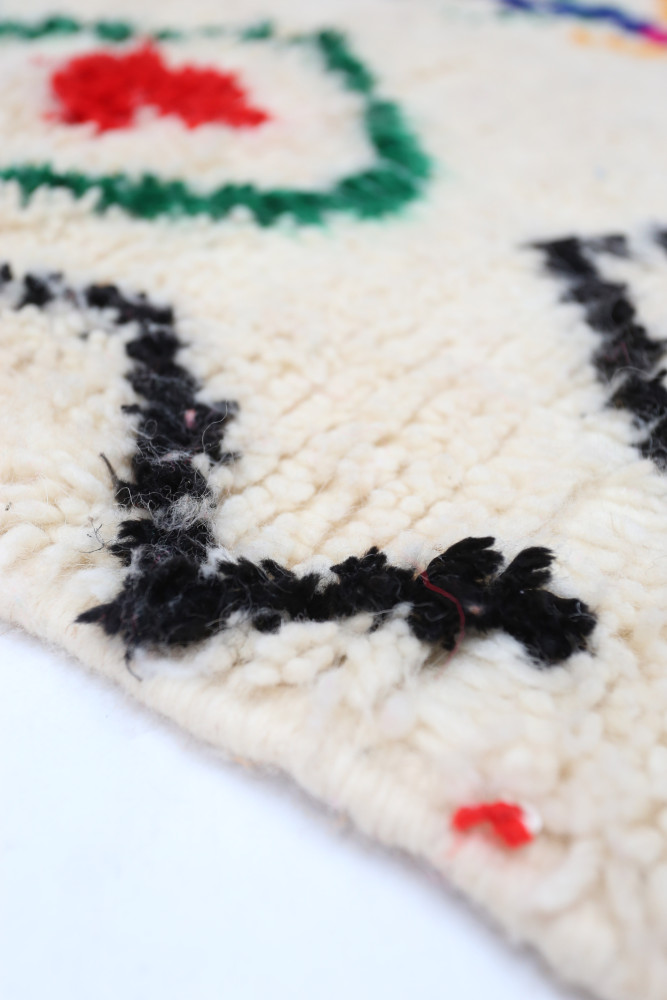 Tapis berbère en laine beige avec motifs géométriques noirs, rouges et verts, ajoutant une touche artisanale à votre intérieur.