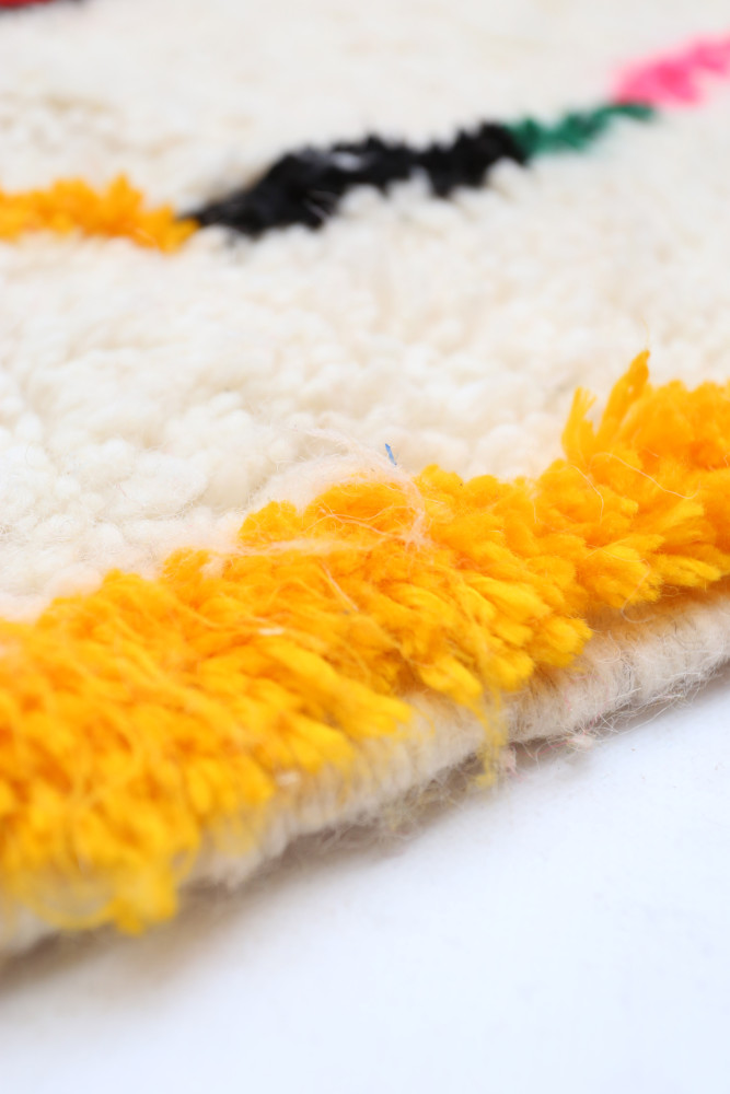 Tapis berbère aux motifs géométriques, couleurs vives, dominé par le blanc, noir et des touches de jaune, rose et vert.