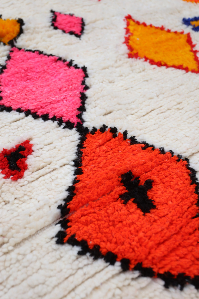 Tapis berbère aux motifs géométriques colorés en rose, orange et noir sur un fond crème.