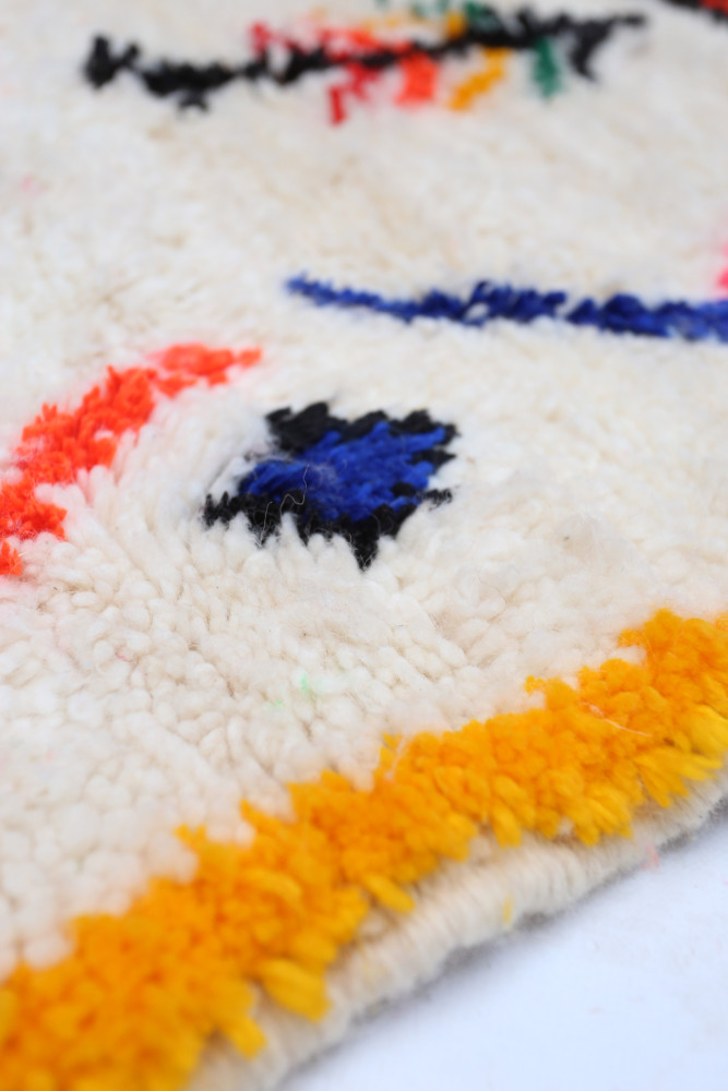 Tapis berbère aux motifs colorés avec des tons blancs, noirs, orange et jaune, apportant une touche artisanale et chaleureuse.