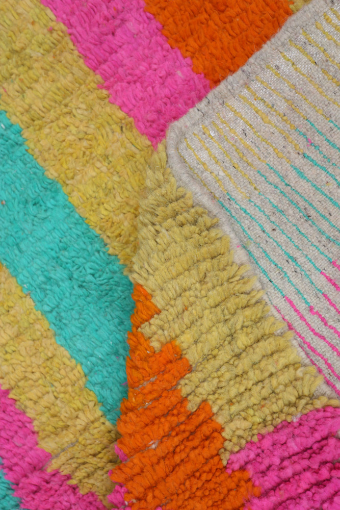 Tapis berbère coloré avec des motifs géométriques vifs en rose, orange, turquoise et jaune sur fond texturé.