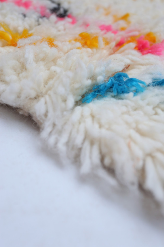 Tapis berbère en laine blanche avec des motifs colorés rose, bleu et jaune, offrant un style artisanal et chaleureux.