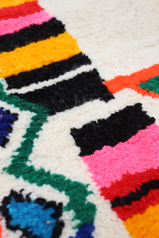 Tapis berbère coloré avec des motifs géométriques en noir, rose, orange et vert sur un fond crème.