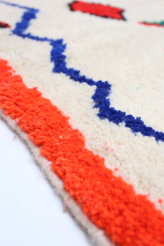 Tapis berbère avec des motifs géométriques colorés en bleu, rouge, et orange sur un fond crème.