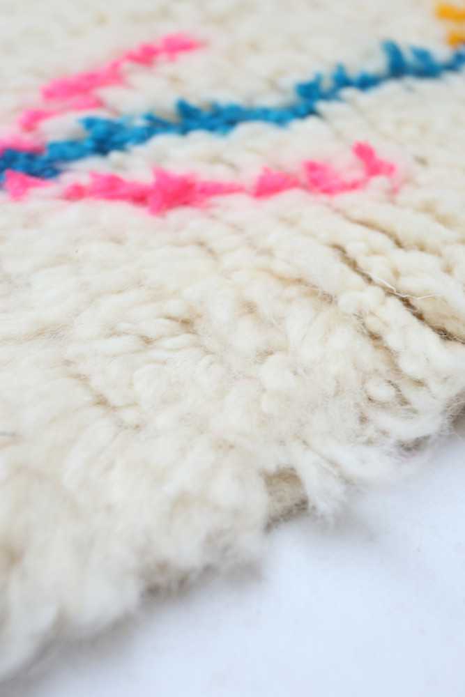 Tapis berbère en laine, avec des motifs colorés en rose, bleu et jaune sur un fond crème.