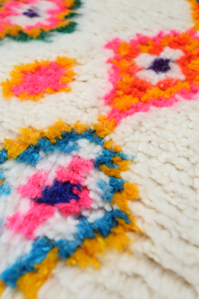 Tapis berbère aux motifs colorés avec des fleurs en rose, orange, bleu et blanc sur fond crème.