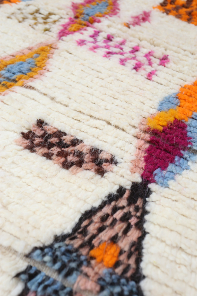 Tapis berbère aux motifs colorés avec des teintes vives de bleu, rose, orange et marron sur fond blanc.
