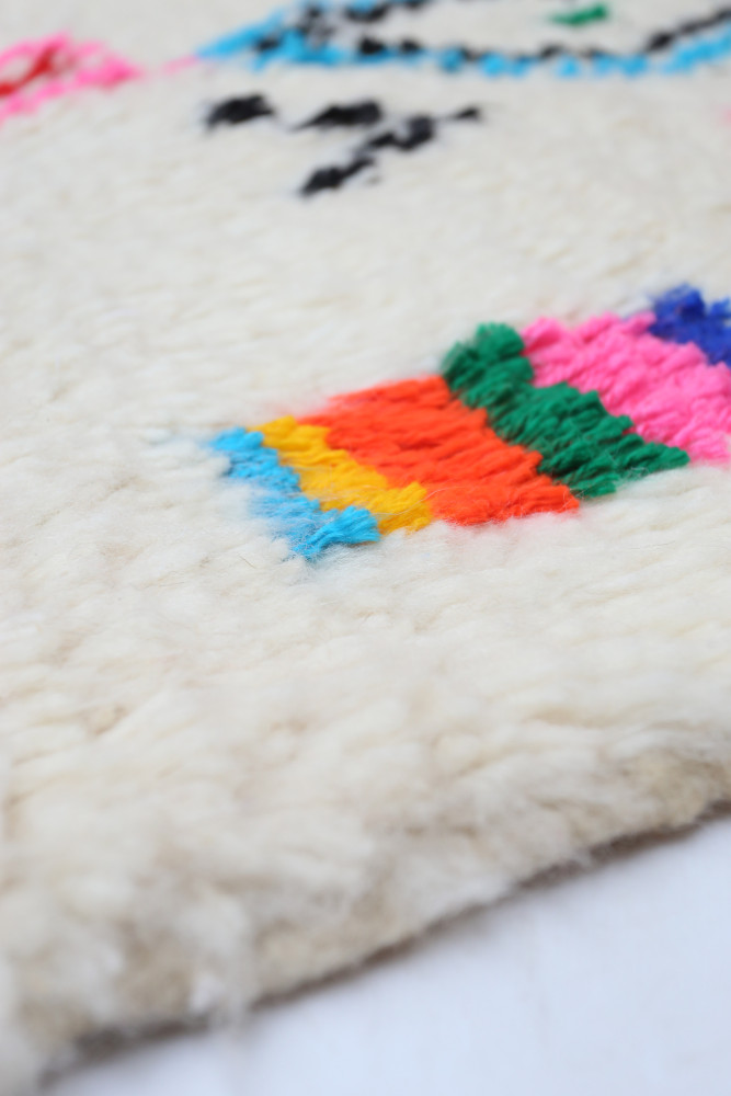 Tapis berbère en laine avec motifs géométriques colorés sur fond crème et touches de rose, bleu, vert et orange.
