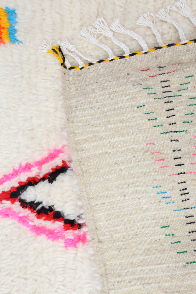 Tapis berbère aux motifs géométriques colorés en rose, noir, bleu et vert sur un fond crémeux.