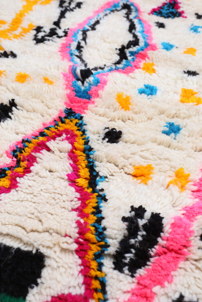 Tapis berbère coloré, aux motifs géométriques avec des tons blancs, roses, noirs, et jaunes, ajoutant une touche bohème.