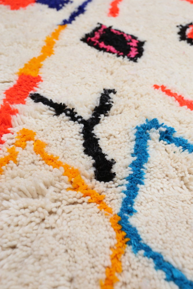 Tapis berbère coloré avec des motifs géométriques noirs, bleus, oranges et roses sur un fond crème.