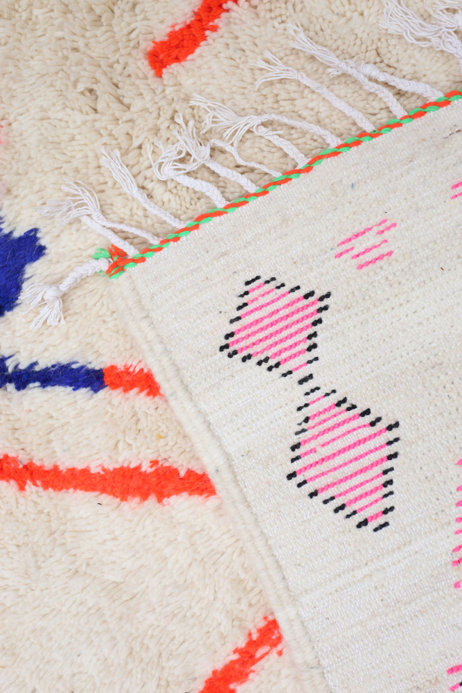 Tapis berbère en laine naturelle, aux motifs géométriques colorés de rose, bleu et orange sur un fond crème.