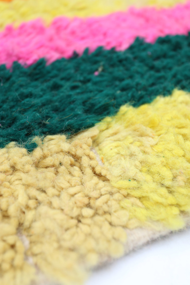 Tapis berbère coloré avec des rayures vibrantes de rose, vert, jaune et beige, présentant un motif texturé et modulaire.
