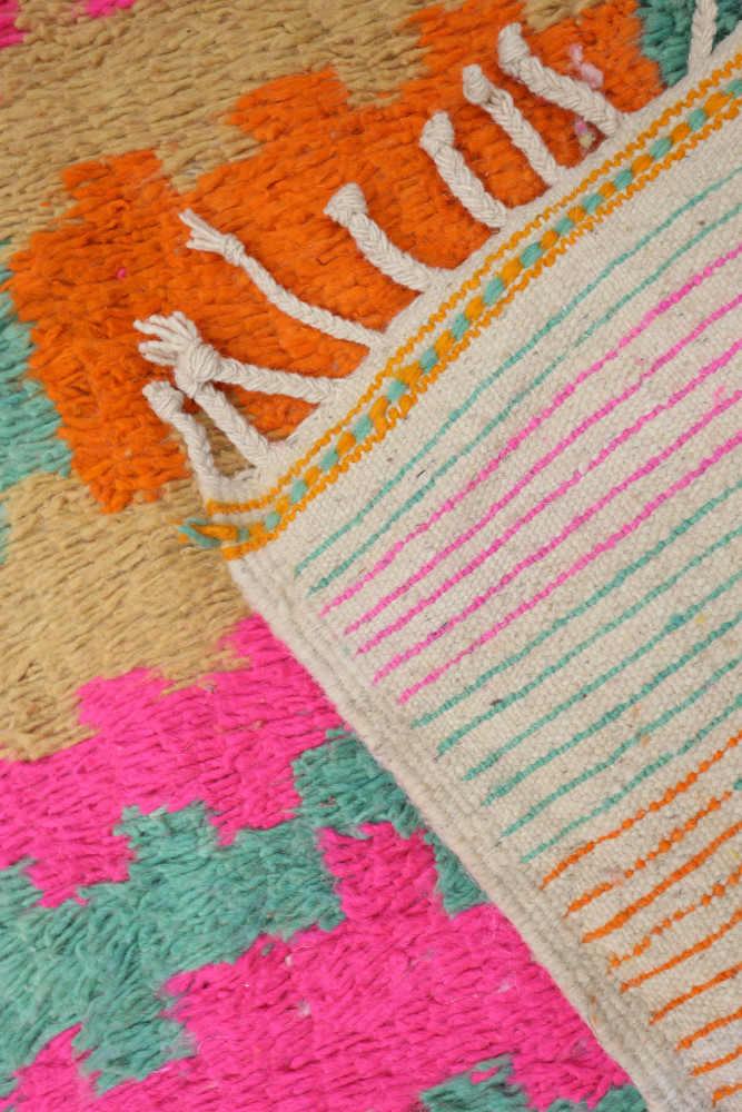 Tapis berbère aux motifs géométriques colorés, mélange de rose, orange, et vert sur fond crème avec des franges décoratives.