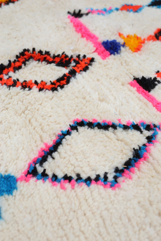 Tapis berbère en laine avec un fond crème et des motifs géométriques colorés en rose, noir, bleu et orange.