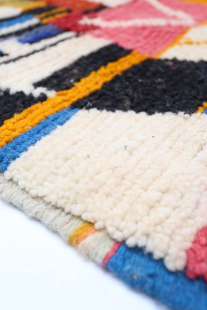 Tapis berbère coloré avec des motifs géométriques en rose, noir, jaune et bleu sur un fond crémeux.