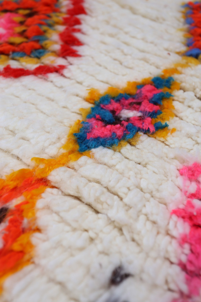 Tapis berbère aux motifs colorés en rose, orange, bleu, sur fond blanc, ajoutant une touche ethnologique à votre intérieur.