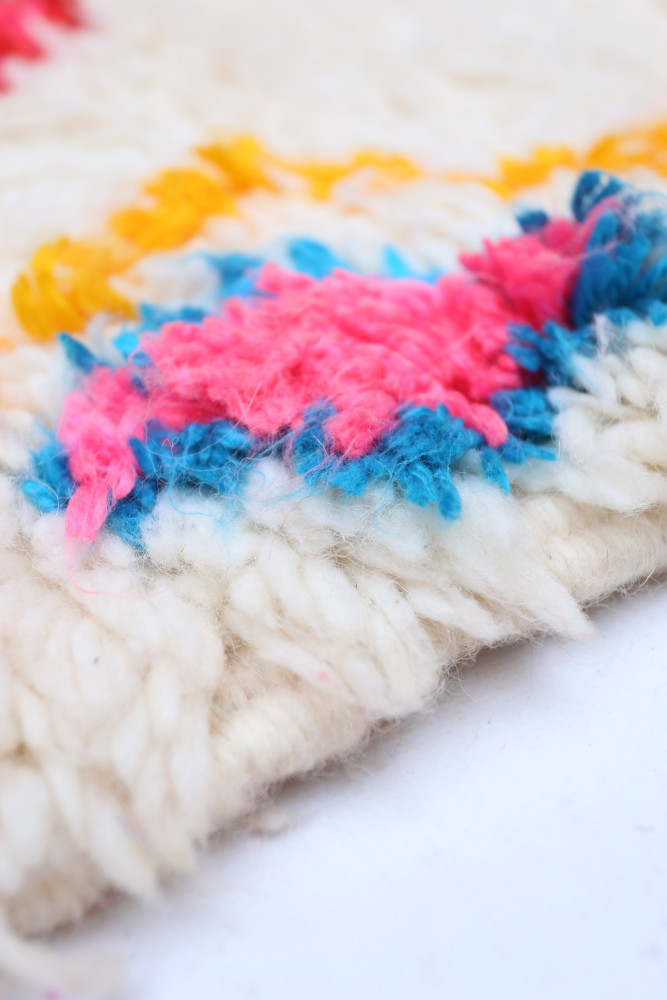 Tapis berbère coloré aux motifs géométriques dans des tons de rose, bleu et jaune sur fond blanc.