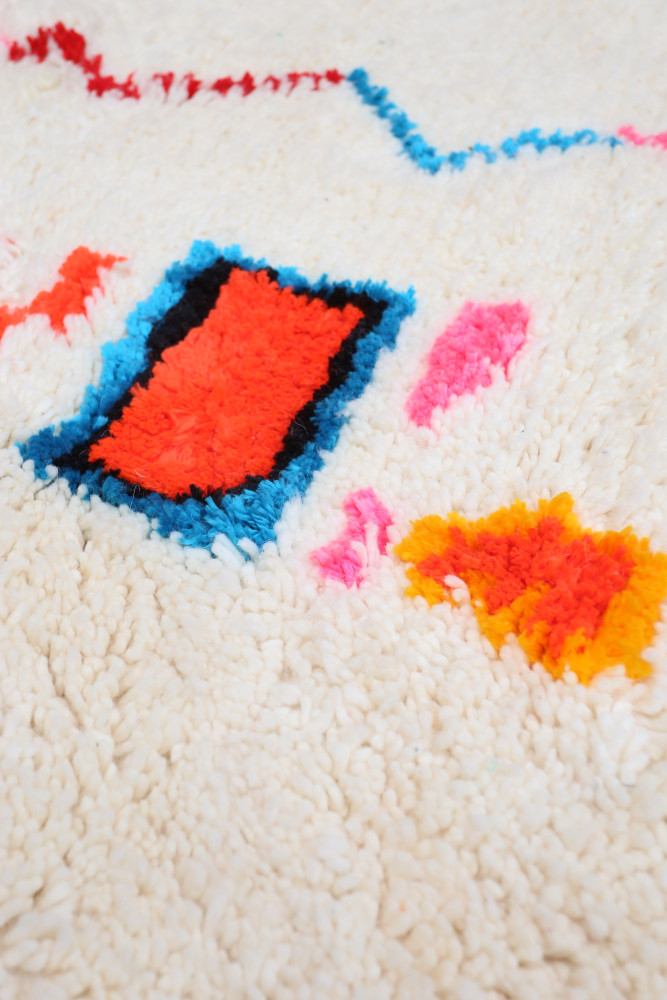 Tapis berbère en laine, aux motifs géométriques colorés de rouge, bleu, rose et orange sur fond crème.