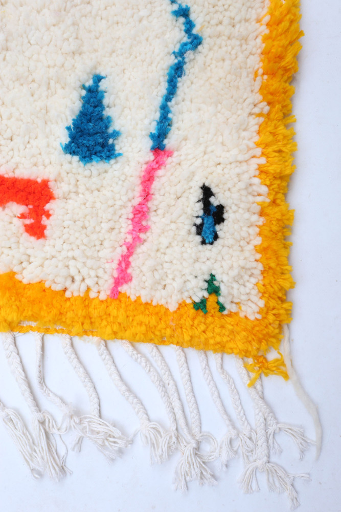 Tapis berbère aux motifs géométriques colorés, mélange de blanc, bleu, rose et orange, bordure frangée joyeuse.