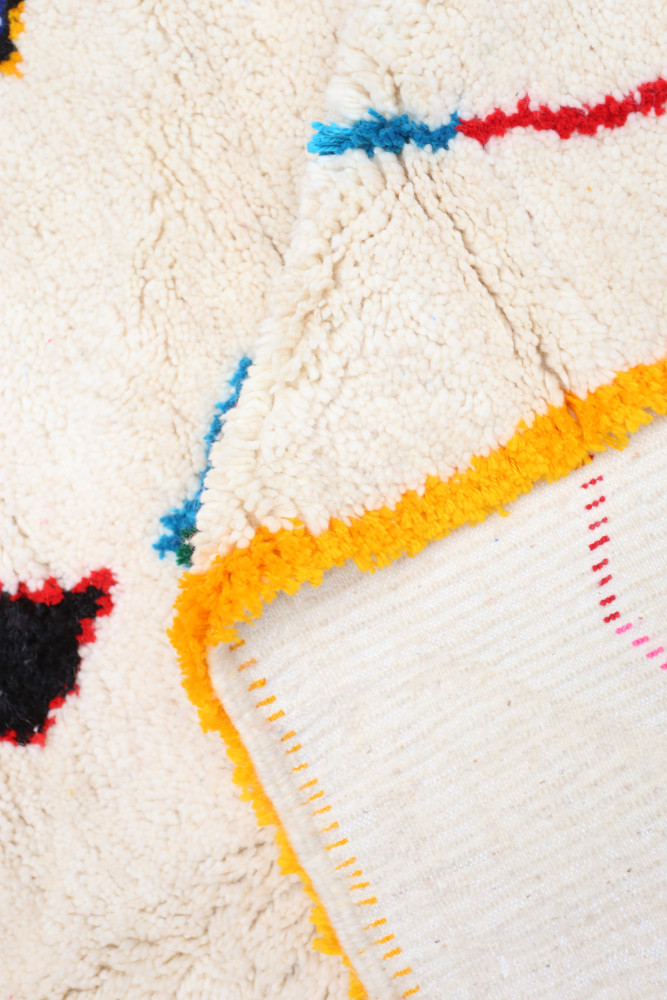 Tapis berbère en laine, crème avec des motifs colorés en bleu, rouge et noir, bordé d\'un liseré orange.