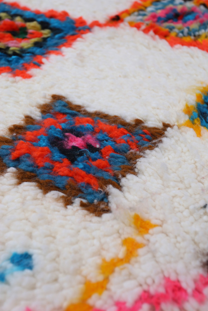 Tapis berbère coloré aux motifs géométriques vibrants sur un fond crème, mélange de bleus, rouges et roses.