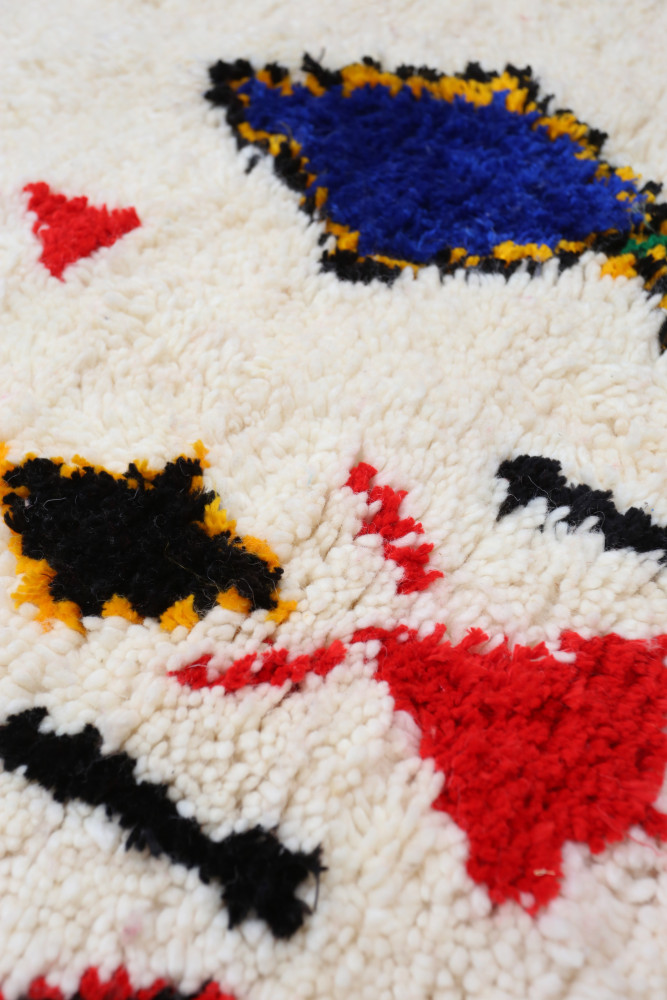 Tapis berbère au style coloré, avec des motifs géométriques en bleu, rouge, noir et jaune sur fond beige.