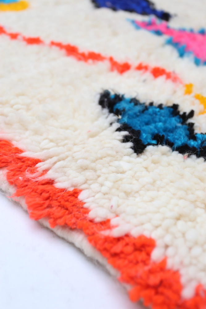 Tapis berbère doux à motifs géométriques colorés sur fond crème avec des touches de rose, bleu et noir.
