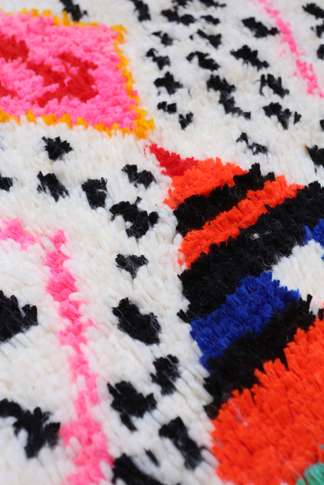 Tapis berbère aux motifs géométriques colorés, avec des tons de blanc, noir, rose, orange et bleu.