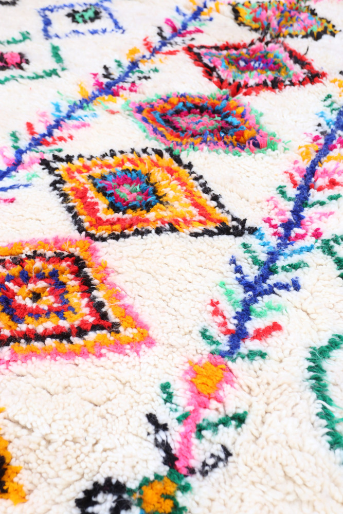 Tapis berbère aux motifs colorés en losanges orange, bleu et rose sur un fond crémeux et texturé.