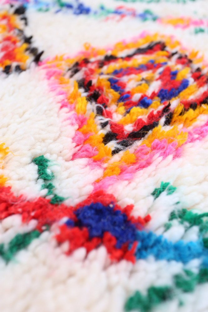 Tapis berbère coloré avec des motifs géométriques en rouge, bleu, jaune, et rose sur un fond crémeux.