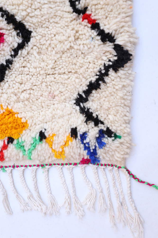 Tapis berbère aux motifs géométriques noirs sur fond crème, avec des touches de couleurs vives et des franges.