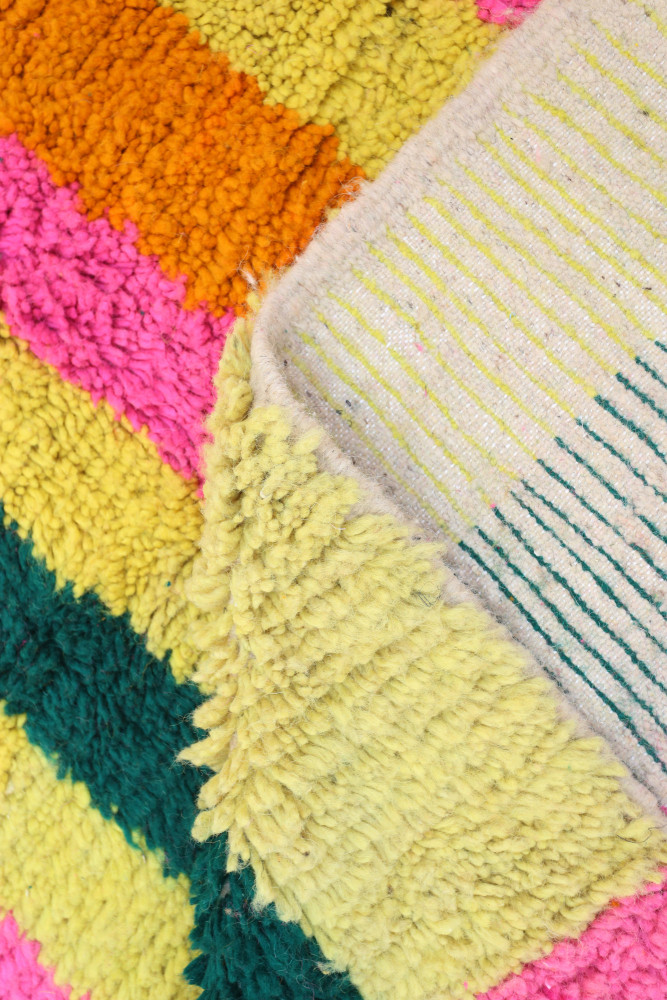 Tapis berbère coloré avec des motifs géométriques en rose, orange, vert et jaune sur un fond texturé.