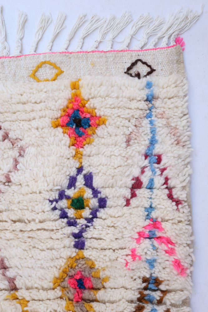 Tapis berbère en laine écrue ornée de motifs géométriques colorés comme le rose, le bleu, le jaune et le violet.