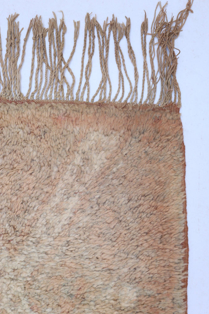Tapis berbère artisanal aux teintes beige et rose, orné de motifs géométriques et franges délicates.